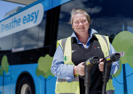 Smiling female bus driver holding charging cables in front of a blue zero-emission bus.